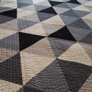 May include: A geometric quilt with a repeating triangle pattern in black, grey, and cream. The quilt has a textured surface with stitched lines, creating a modern design. This bedding item is suitable for a bed or as a decorative throw, adding a stylish touch.