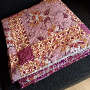 Puede incluir: Un edred&oacute;n plegado con un patr&oacute;n floral y geom&eacute;trico en tonos rosa, marr&oacute;n y morado. El edred&oacute;n presenta un dise&ntilde;o acolchado y est&aacute; doblado cuidadosamente, mostrando su intrincado patchwork y paleta de colores.