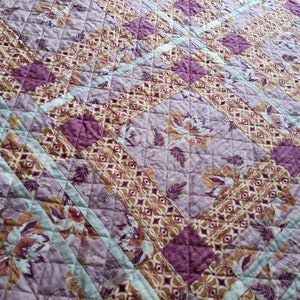 Puede incluir: Un edred&oacute;n acolchado con un estampado floral en tonos morado, marr&oacute;n y crema. El dise&ntilde;o presenta formas geom&eacute;tricas repetidas y motivos florales, con rayas de acento azul claro. El edred&oacute;n est&aacute; hecho de tela suave.