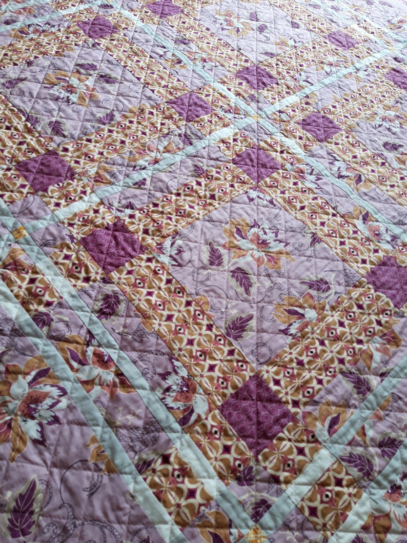 Puede incluir: Un edred&oacute;n acolchado con un estampado floral en tonos morado, marr&oacute;n y crema. El dise&ntilde;o presenta formas geom&eacute;tricas repetidas y motivos florales, con rayas de acento azul claro. El edred&oacute;n est&aacute; hecho de tela suave.