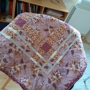 Puede incluir: Una cubierta de mesa redonda acolchada con un estampado floral en tonos rosa, marr&oacute;n y morado. La cubierta presenta un patr&oacute;n de diamantes con un borde gris claro y un borde morado oscuro. El dise&ntilde;o floral incluye flores y hojas grandes y estilizadas.