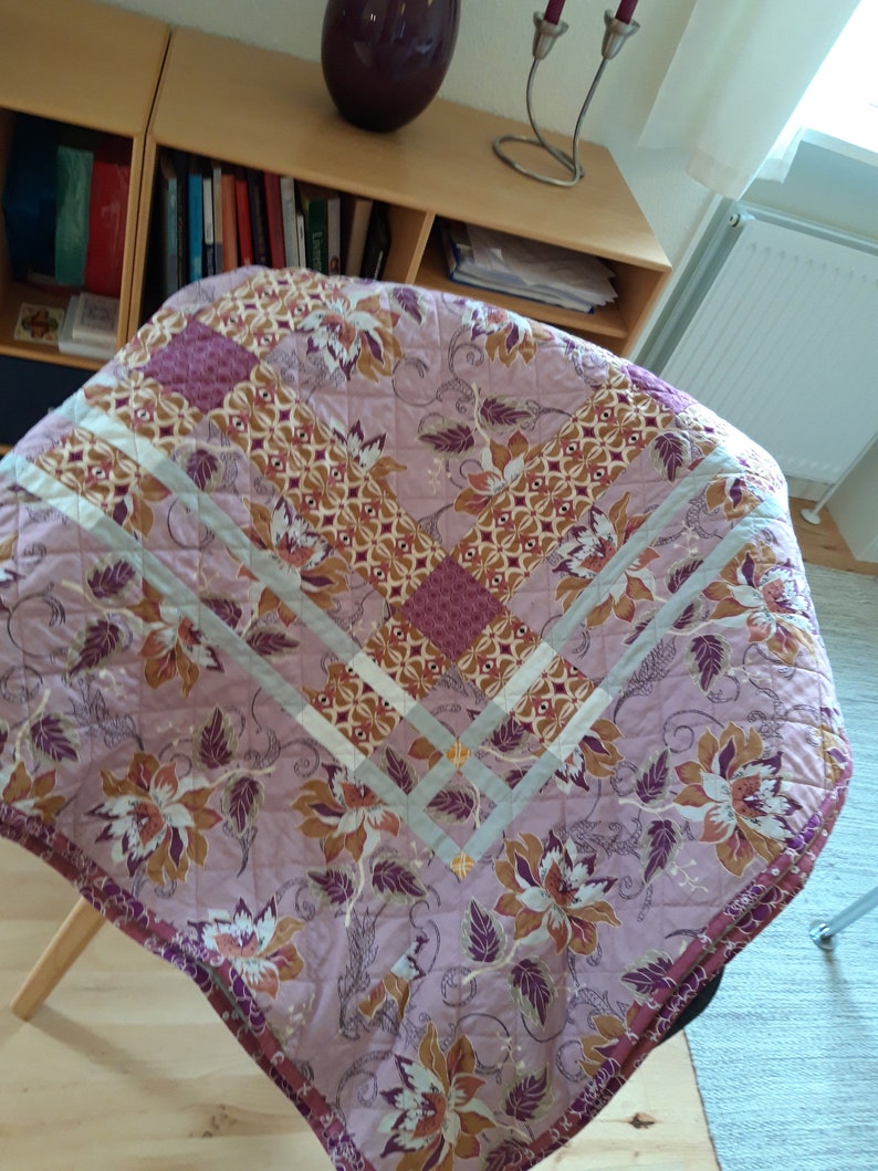 Puede incluir: Una cubierta de mesa redonda acolchada con un estampado floral en tonos rosa, marr&oacute;n y morado. La cubierta presenta un patr&oacute;n de diamantes con un borde gris claro y un borde morado oscuro. El dise&ntilde;o floral incluye flores y hojas grandes y estilizadas.