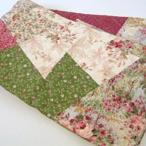 Op de afbeelding: Een opgevouwen quilt met een patchwork-ontwerp. De quilt heeft verschillende bloempatronen in tinten roze, groen en crème. De quilt is gewatteerd met een ruitpatroon. De quilt is opgevouwen en lijkt van katoen te zijn.