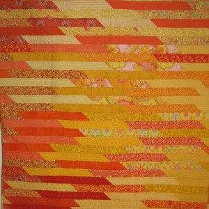 May include: A handmade quilt with a vibrant, warm color palette. The quilt features horizontal bands of various shades of yellow, orange, and red fabrics. The design incorporates diagonal, geometric patterns, creating a visually dynamic textile.