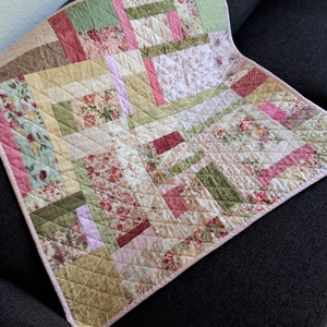 May include: A handmade quilt with a patchwork design. The quilt features floral and geometric patterns in pink, green, and cream. The quilt measures approximately 100 cm by 100 cm, with a pink border.