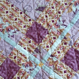 Puede incluir: Un edred&oacute;n de patchwork con un dise&ntilde;o floral y geom&eacute;trico. El patr&oacute;n del edred&oacute;n incluye formas en lavanda, marr&oacute;n y burdeos. Tiras de color azul claro se cruzan, creando un efecto de cuadr&iacute;cula. La tela est&aacute; cosida.