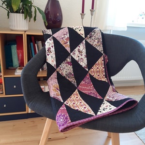 May include: A handmade quilt draped over a gray chair. The quilt features a geometric pattern of black triangles interspersed with floral print triangles in shades of pink, purple, and cream. The quilt's edge is trimmed with a solid purple border.