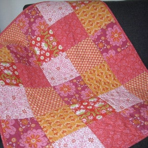 Puede incluir: Un edredón de patchwork con patrones florales y geométricos en tonos rosa, naranja y rojo. El edredón está hecho de cuadrados cosidos, creando un diseño visualmente atractivo. El edredón probablemente está hecho de algodón.