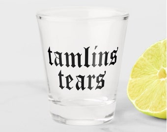 May include: A clear shot glass with the words "tamlin's tears" in black gothic font. A slice of lime is visible to the right of the glass. The shot glass is on a white surface.