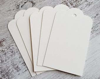 May include: A stack of six blank white gift tags with scalloped edges and a hole for string. The tags are on a light gray wooden surface with small red hearts scattered around.