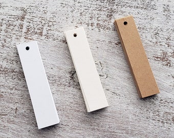 May include: Three rectangular paper tags with holes punched at the top. The tags are white, off-white, and brown. The tags are arranged horizontally on a light wood surface.