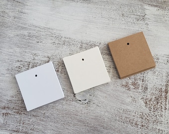May include: Three square blank tags in white, off-white, and brown, each with a small hole at the top. The tags are arranged diagonally on a distressed, white-painted wooden surface.