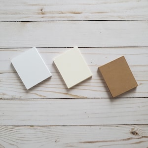May include: Three square paper notepads in different colors: white, cream, and brown. The notepads are arranged in a row on a white wooden surface.