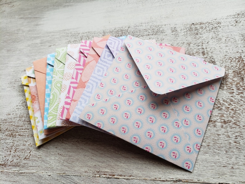 May include: A stack of colorful patterned envelopes with a light blue envelope on top. The envelopes are folded and have a variety of floral and geometric designs.