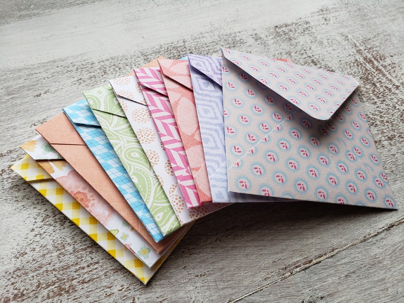 May include: A stack of ten colorful patterned envelopes with different designs. The envelopes are folded and stacked on top of each other, with the top envelope being a light blue with a white and pink floral pattern.