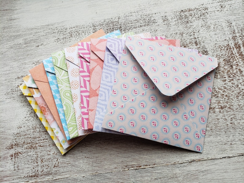 May include: A stack of colorful patterned envelopes with a variety of designs. The envelopes are folded and stacked on top of each other, with the top envelope being a light pink with a small blue and pink floral pattern.