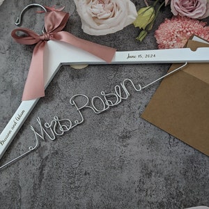 May include: A white wooden wedding dress hanger with a pink satin ribbon and the names "Breann and Adam" engraved on the side. The date "June 15, 2024" is printed on the top. A wire "Mrs. Rosen" sign and brown envelopes are also present.