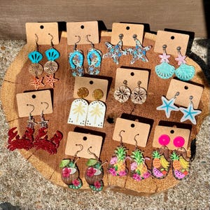 May include: A collection of twelve pairs of summer-themed dangle earrings displayed on brown cardboard cards. Designs include crabs, flip-flops, starfish, palm trees, pineapples, tropical drinks, seashells, and fruit slices in various bright colors.