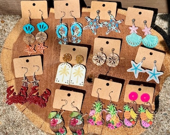 May include: A collection of twelve pairs of summer-themed dangle earrings displayed on brown cardboard cards. Designs include crabs, flip-flops, starfish, palm trees, pineapples, tropical drinks, seashells, and fruit slices in various bright colors.