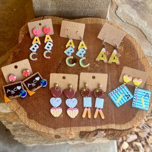 May include: A selection of colourful acrylic earrings with school-themed designs. Styles include apple and ABC letter earrings, chalkboard earrings, heart and pencil earrings, and notebook earrings. The earrings are displayed on brown cardboard earring cards.
