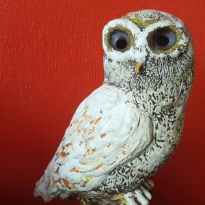 Può includere: Una statuetta di gufo in ceramica con finitura bianca testurizzata, dettagli neri incisi e sottili accenti arancioni sulle ali. Il gufo presenta grandi occhi scuri e cavi e un piccolo becco giallo, raffigurato in posizione eretta.