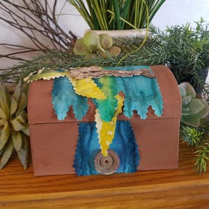 May include: A small, brown wooden box with a hinged lid. The box is decorated with blue and yellow fabric scraps and a brown button.