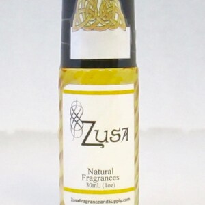 May include: A clear glass roll-on bottle with a black cap featuring a gold Celtic knot design. The label reads "Zusa Natural Fragrances 30ml (1oz)" with a stylized logo. The bottle contains a yellow liquid.