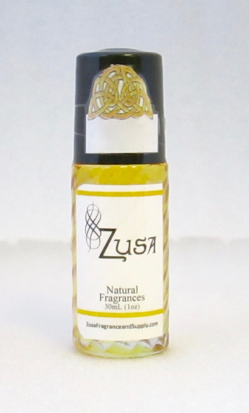 May include: A clear glass roll-on bottle with a black cap featuring a gold Celtic knot design. The label reads "Zusa Natural Fragrances 30ml (1oz)" with a stylized logo. The bottle contains a yellow liquid.