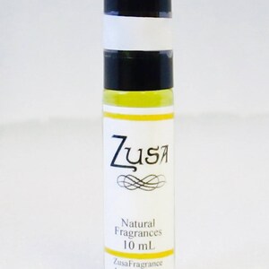 May include: A clear glass roll-on bottle with a black cap and a white label. The label features the word "Zusa" in a stylized font, with "Natural Fragrances" and "10 mL" below. The website ZusaFragranceAndSupply.com is also printed on the label.