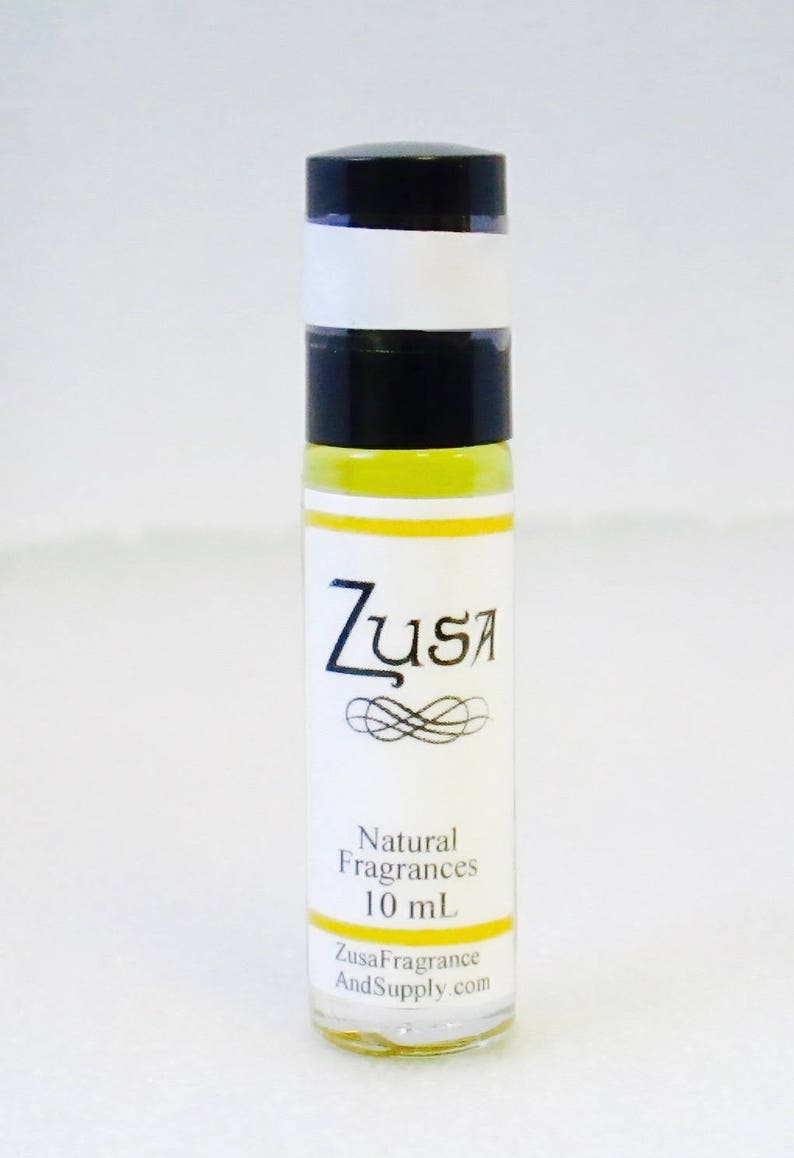May include: A clear glass roll-on bottle with a black cap and a white label. The label features the word "Zusa" in a stylized font, with "Natural Fragrances" and "10 mL" below. The website ZusaFragranceAndSupply.com is also printed on the label.