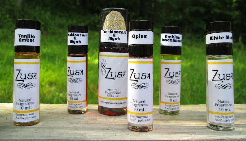 May include: A collection of Zusa Natural Fragrances in various scents, including Vanilla Amber, Frankincense & Myrrh, Opium, Arabian Sandalwood, and White Musk. Each 10 mL bottle has a white label with black text and a yellow stripe.