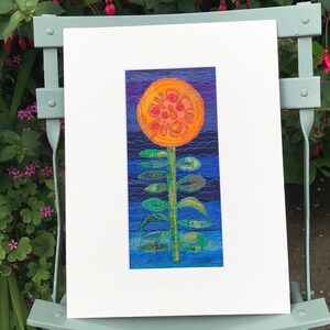 May include: A framed textile art piece featuring a stylised orange flower with a green stem and leaves against a blue and purple background.