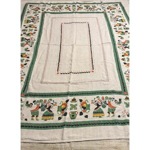 May include: A rectangular beige tablecloth featuring a decorative border with repeating illustrations of figures in colorful attire, floral arrangements, and roosters. The center is framed by a thin black and red geometric stitched pattern.