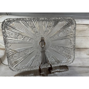 Könnte beinhalten: Ein klarer, rechteckiger Servierteller aus Glas mit komplizierten Sternen- und geometrischen Mustern. Das Tablett hat einen dekorativen Rand und einen zentralen Griff. Es wird auf einem bronzefarbenen Ständer präsentiert, der sein elegantes Design zeigt.