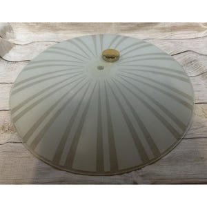 Könnte beinhalten: Eine runde, vintage Deckenleuchte aus Milchglas mit einem Sonnenstrahl-Design. Die Leuchte hat einen messingfarbenen Mittelknopf. Das Glas hat ein weißes und graues Farbschema. Die Leuchte ist ein Einrichtungsgegenstand.