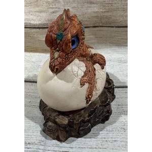 Peut inclure: Figurine détaillée d'un bébé dragon sortant d'un œuf. Le dragon est brun avec des yeux bleus et des bijoux décoratifs. L'œuf est blanc cassé et la base est une formation rocheuse.