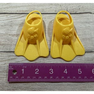 May include: A pair of bright yellow toy swim fins. Each fin has a loop for the foot and a fish design. The fins are approximately 4 inches long. The fins are on a wooden surface.