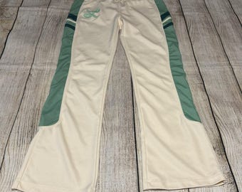 以下が含まれることがあります： ドローストリングウエストとワイドレッグが特徴のクリーム色のトラックパンツ。サイドにはセージグリーンのパネルとダークグリーンのストライプがあしらわれています。右腰部分にはライトグリーンの刺繍ロゴが施されています。