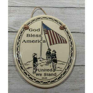 May include: Oval ceramic plaque with the text "God Bless America" and "United We Stand". The plaque features an American flag and illustrations of firefighters. It has a decorative border and a jute string for hanging.