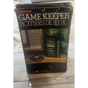 May include: A black Dynasound Game Keeper storage box for video game cartridges. The box features text reading GAME KEEPER BOITIERS DE JEUX and displays a graphic of a cartridge holder. A metal ruler shows the box is 8 inches tall.