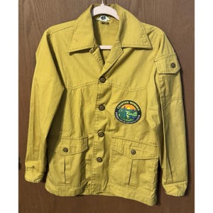 Może przedstawiać: Musztardowa kurtka z czterema przednimi kieszeniami i zapięciem na guziki. Kurtka posiada naszywkę "Charter Member Fred Bear Sports Club" na lewej piersi. Kurtka wisi na białym wieszaku.