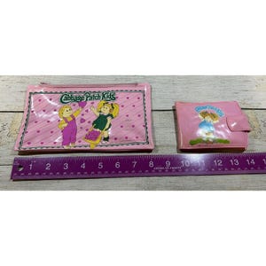 Op de afbeelding: Twee roze Cabbage Patch Kids-accessoires: een rechthoekig etui met ritssluiting en een portemonnee met twee vouwen. De etui heeft twee cartoonkinderen en de tekst "Cabbage Patch Kids". De portemonnee heeft een cartoonkind en dezelfde tekst. Een liniaal toont de afmetingen in centimeters.
