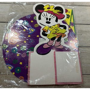 May include: A Minnie Mouse-themed party decoration set. The set includes a die-cut Minnie Mouse figure in a yellow dress and purple shoes, along with a purple circular backdrop with floral and heart designs. The set is packaged in a clear plastic bag.