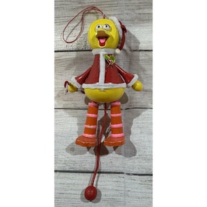 May include: A yellow duck Christmas ornament with a red Santa suit and hat. The ornament has orange and pink striped boots and a red string for hanging. The duck's mouth is open, and it has a holly sprig on its chest.