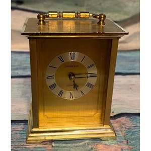 May include: A gold-toned President Quartz clock with Roman numeral hour markers. The clock has a rectangular shape with a handle on top and a gold-colored face. The clock face reads "President" and "Quartz".