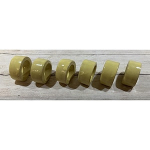 May include: Six ceramic napkin rings in a pale yellow color are arranged on a light wood surface. Each ring is cylindrical with a smooth, glossy finish. The rings are evenly spaced, showcasing their simple, elegant design.