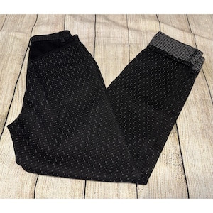 May include: Black pants with a subtle, textured pattern. The pants have a dark waistband and cuffs with a gray and white striped design. The pants are folded on a light wood-grain surface.