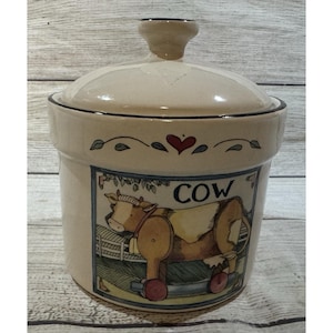 May include: A beige ceramic canister with a lid, featuring a cow illustration and the word "COW". The canister has a black rim and a decorative heart and leaf design. The cow is brown and white.