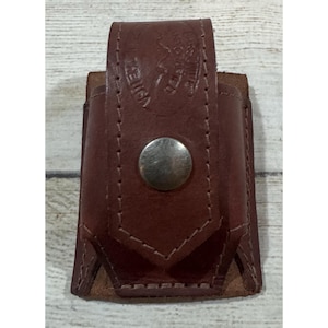 May include: A brown leather pouch with a vertical flap secured by a single silver-toned metal snap button. The leather features visible stitching along the edges and a stamped circular logo on the upper flap that reads Made in the USA.