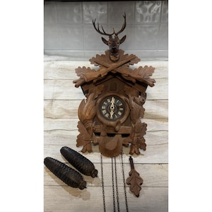 May include: A wooden cuckoo clock with a deer head at the top, carved leaves, and a clock face with Roman numerals. Two pinecone-shaped weights and a leaf-shaped pendulum hang below. The clock is brown.
